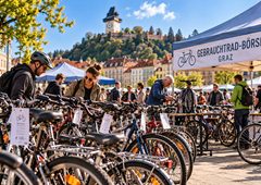 Bildbeschreibung: Redaktionelle Visualisierung einer Fahrrad-Gebrauchtbörse in Graz. Unterschiedliche Fahrräder, Kaufinteressierte und der Grazer Schlossberg veranschaulichen das Thema der historischen BICYCLE-Gebrauchtradbörse und der aktuellen AK-Radbörsen. Wichtiger Bildhinweis: Das Bild wurde mit künstlicher Intelligenz erstellt. Es zeigt keine authentische Aufnahme einer BICYCLE- oder AK-Radbörse. Personen, Verkaufsstände, Beschilderungen und die konkrete Veranstaltungsszene sind fiktiv.
